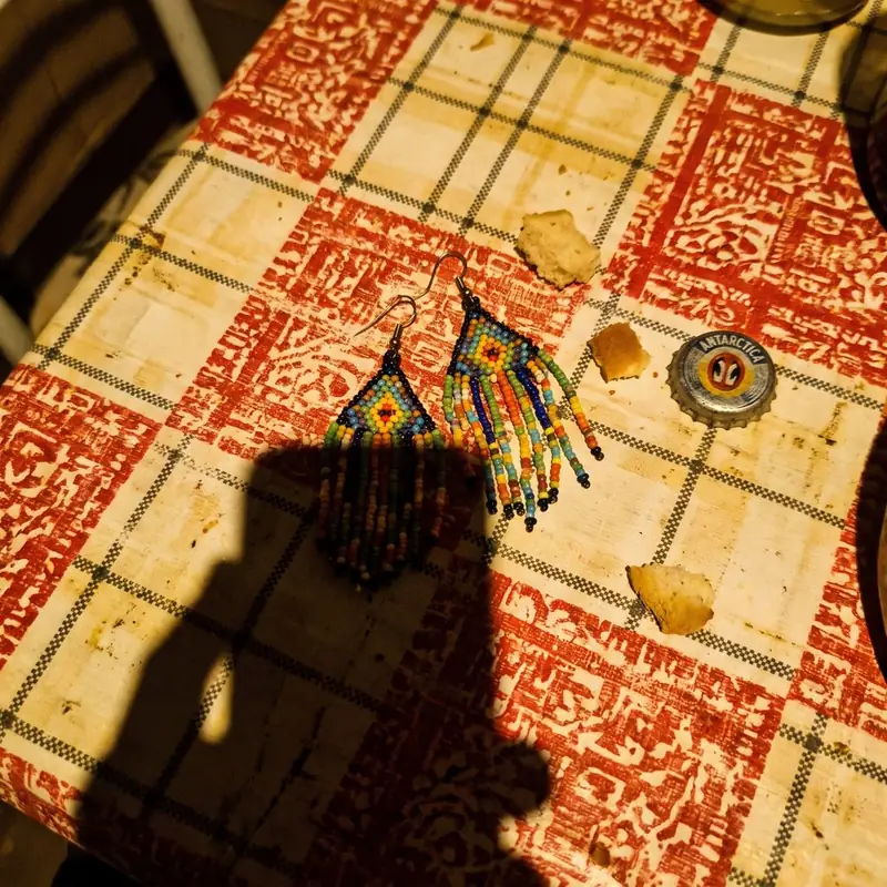 Pair of colorful beaded earrings on a red checkered tablecloth, with crumbs, a bottle cap and the photographer's shadow